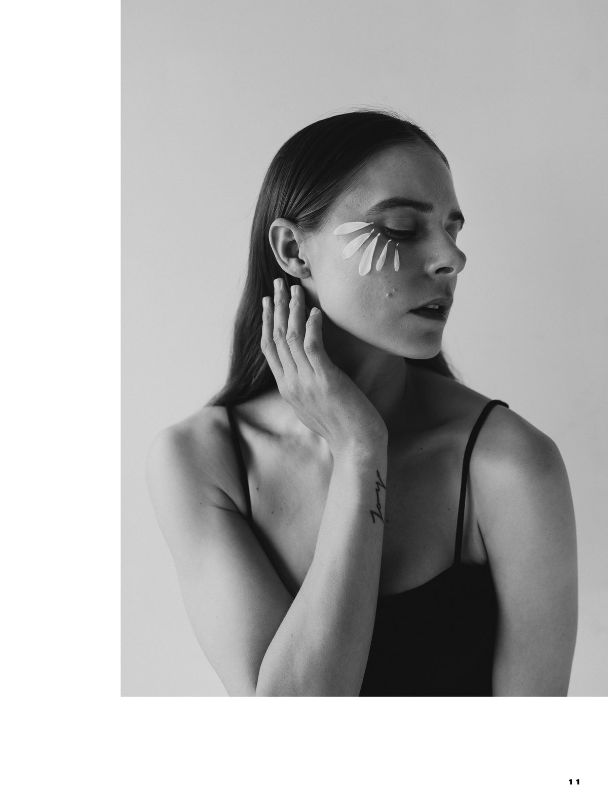Black-and-white portrait of a woman with closed eyes, flower petals arranged beneath one eye and a hand touching her neck.