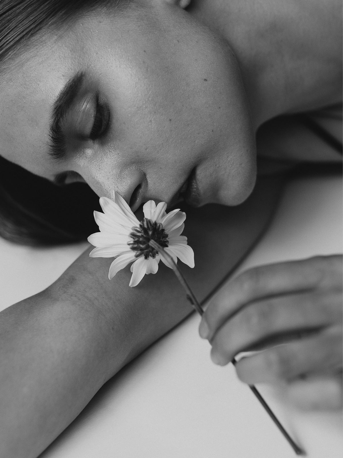 Black-and-white close-up of a reclining woman holding a flower near her nose, her eyes closed.