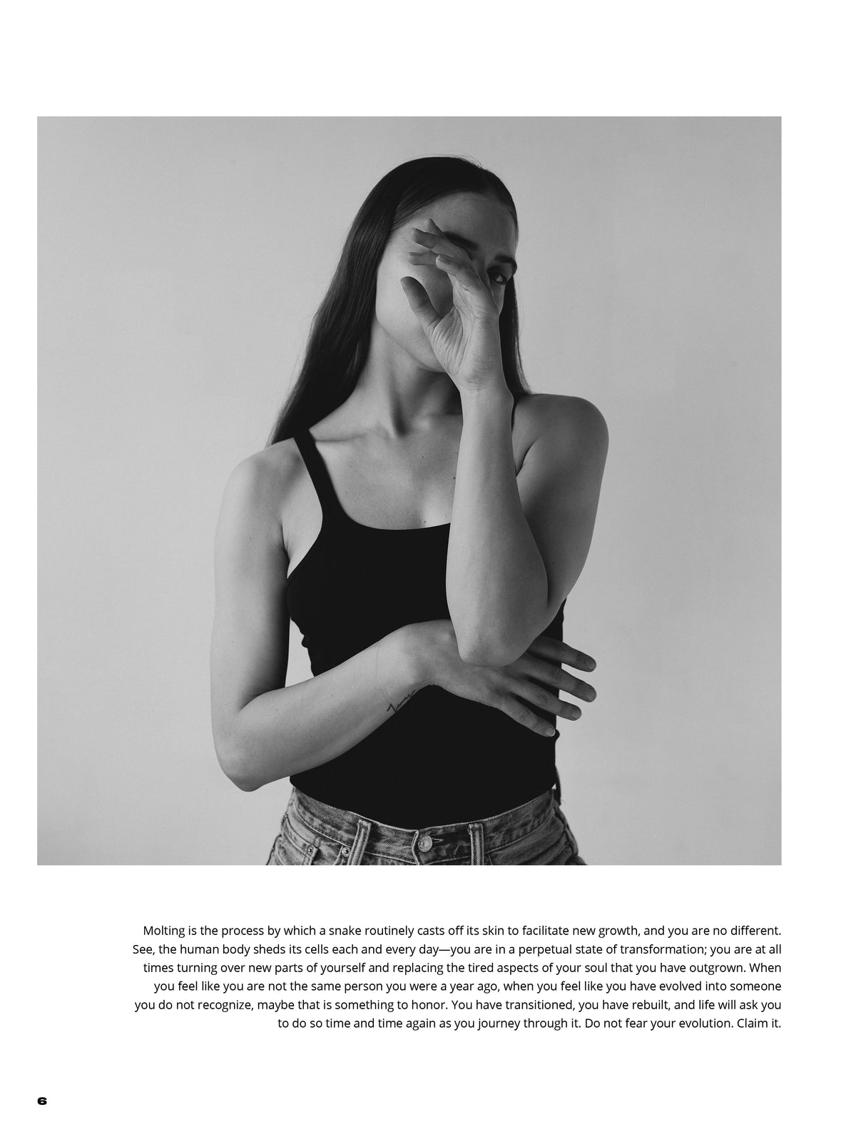 Black-and-white portrait of a woman in a black top and jeans, one arm across her torso and the other hand partly covering her face.
