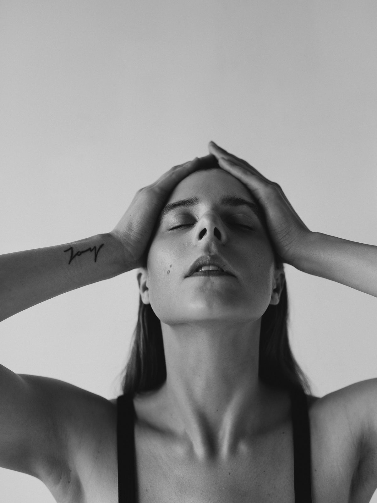 Black-and-white close-up of a woman with closed eyes and both hands pressed to the top of her head.