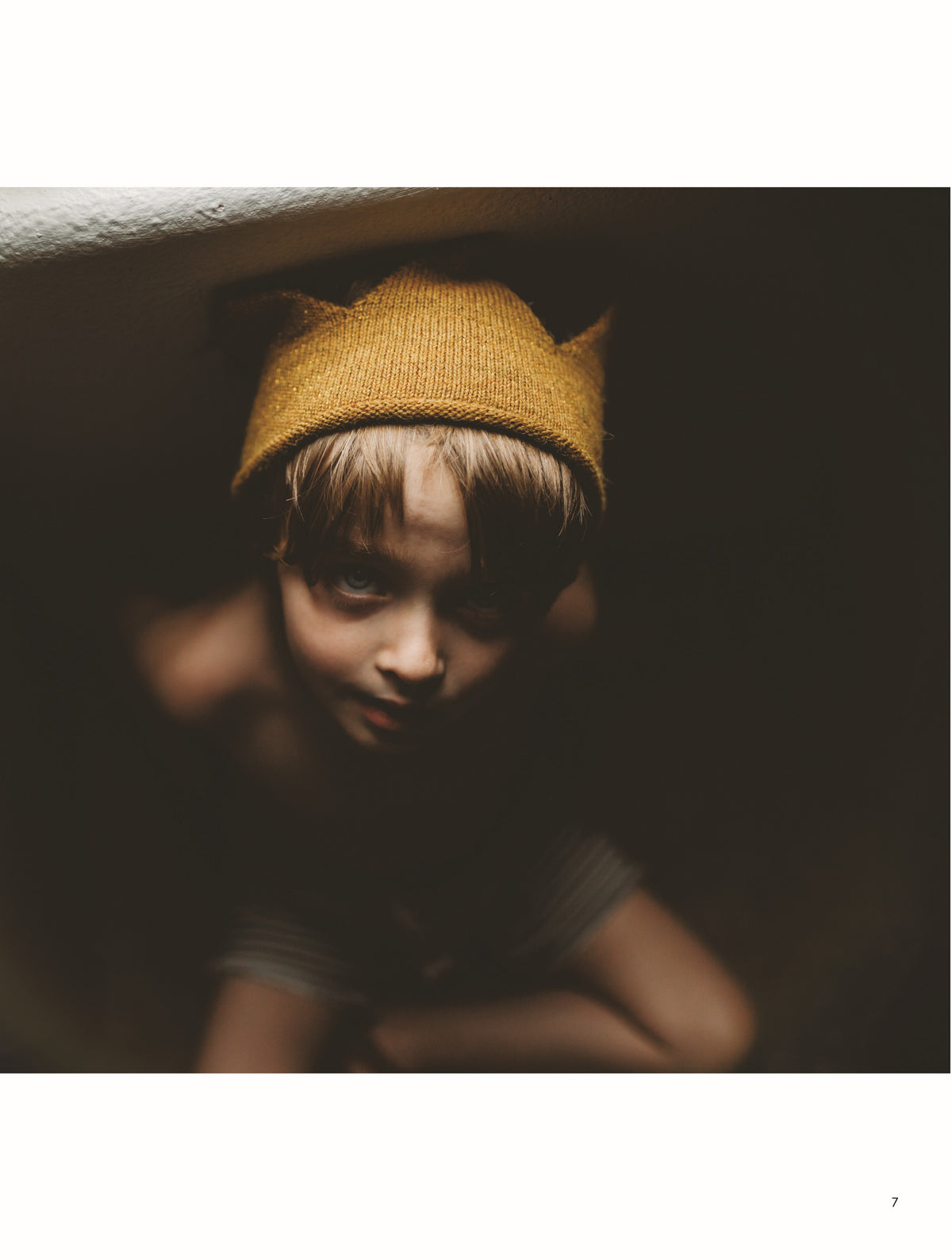 A child wearing a gold knitted crown looks upward toward the camera in a softly lit portrait.