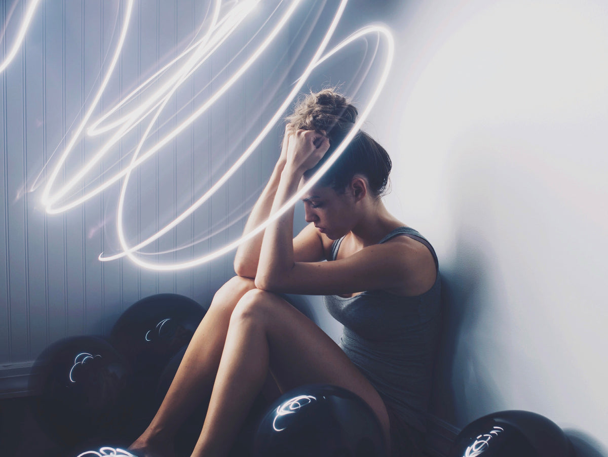 A person sits curled up against a wall among black balloons, head bowed and hands clasped above their head, while bright white light trails loop overhead.