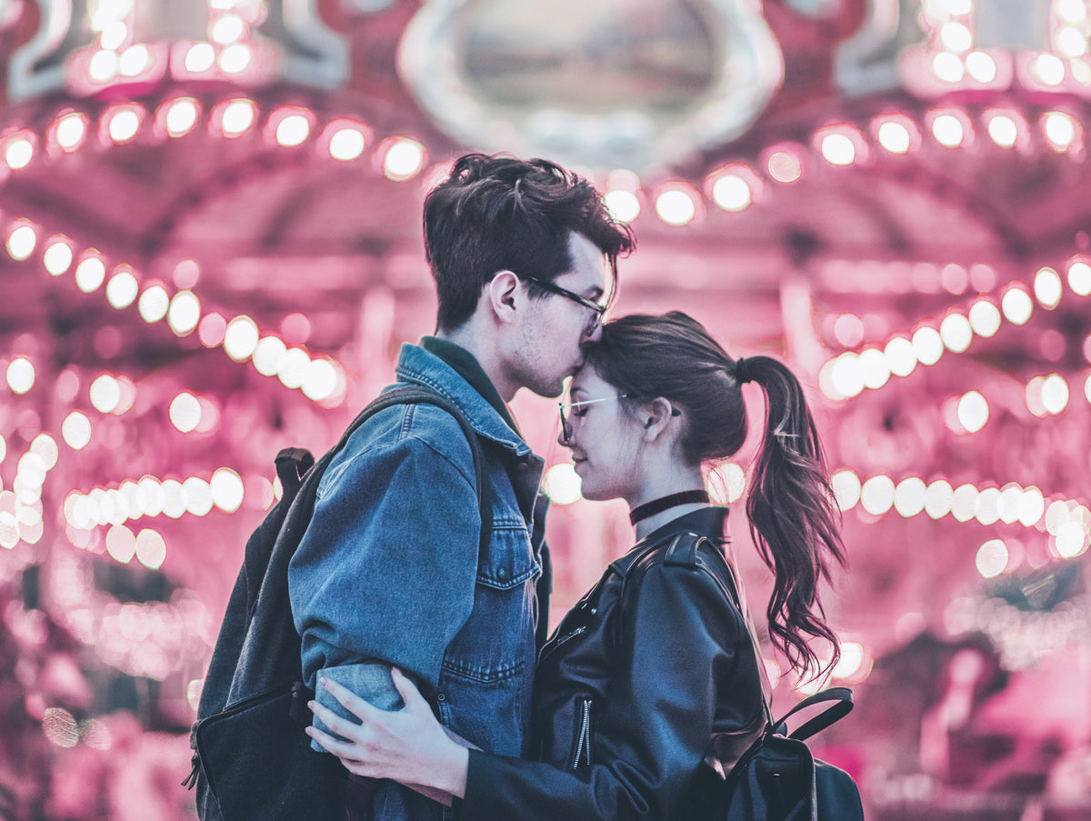 Two people embrace in front of a carousel glowing pink; one kisses the other’s forehead, with strings of blurred bulbs behind them.