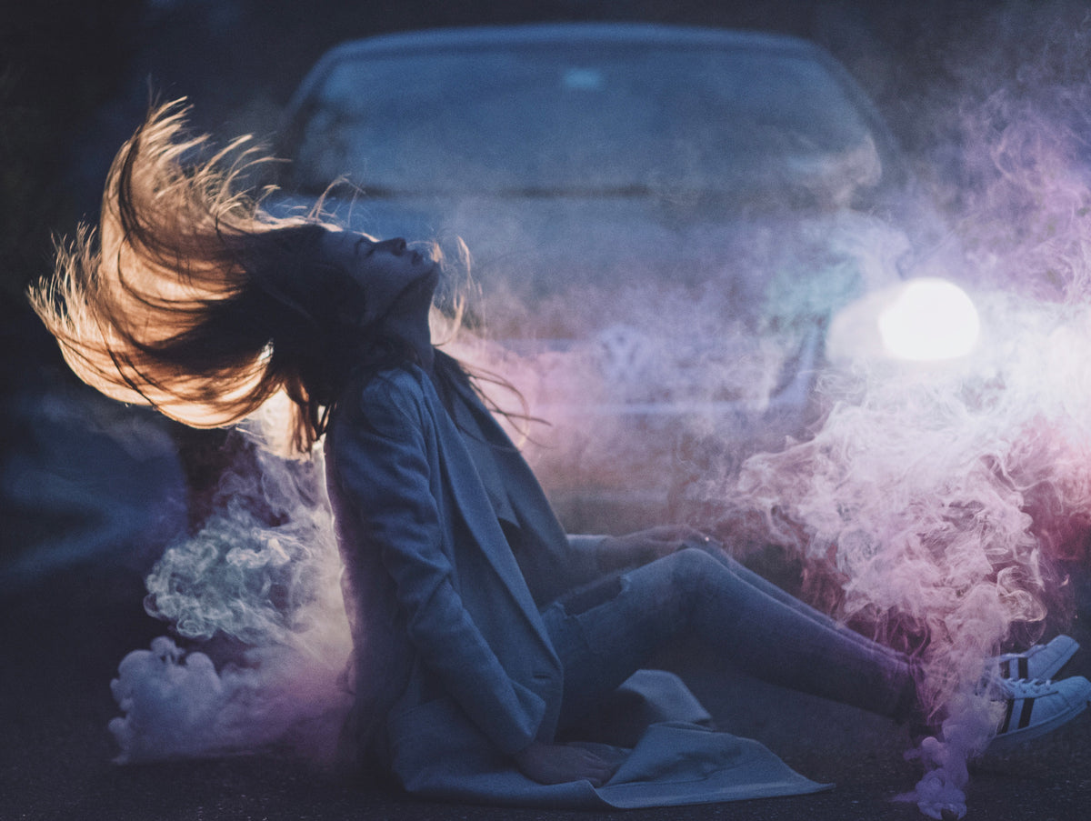 A person sits on the ground and throws their hair back in front of a car, surrounded by pink and purple smoke illuminated by the headlights.